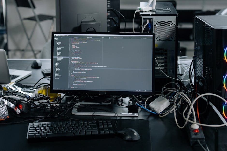 A cluttered workspace featuring a desktop computer with a large monitor displaying lines of code, surrounded by various electronic components and tangled cables. To the left, part of a closed laptop a